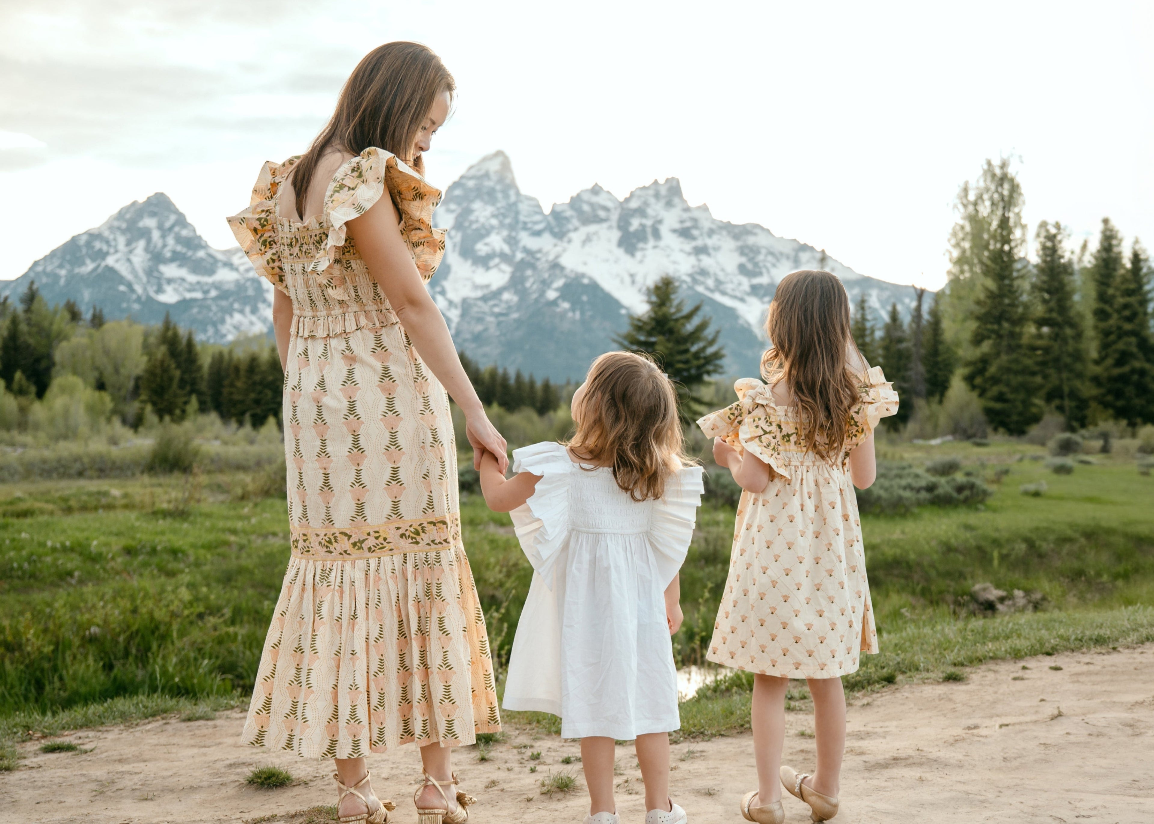Woman and two children in matching dresses walking on a dirt path with mountains in the background
